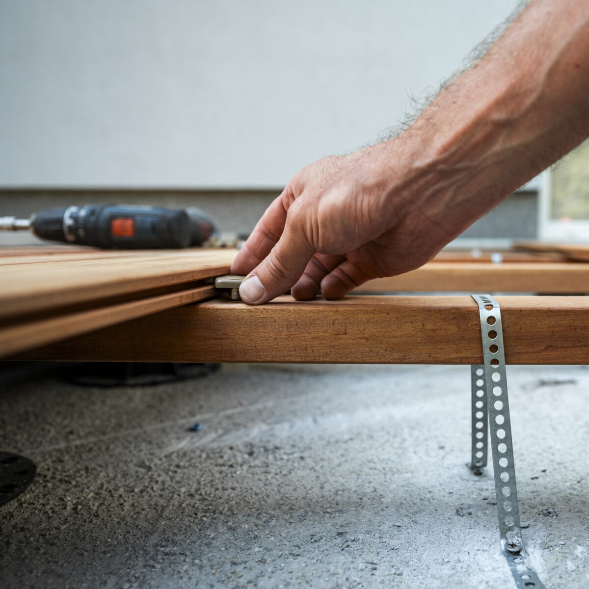 A hand carefully adjusting a wooden board during a construction project of patio, with a power drill in background, highlighting attention to detail and craftsmanship in woodworking.
